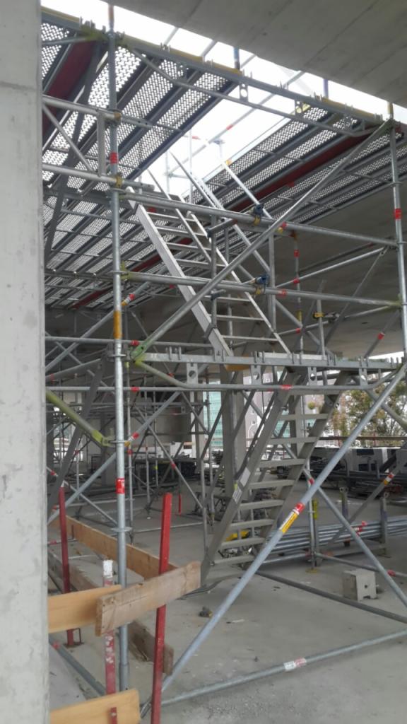 Skomplikowana konstrukcja rusztowania z metalowymi schodami wewnątrz budynku w trakcie budowy, widoczne elementy betonowe i stalowe, oświetlenie dzienne z góry.