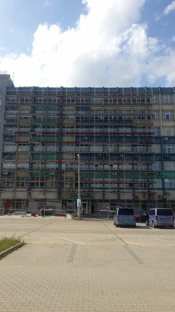 Budynek wielopiętrowy w trakcie renowacji, otoczony rusztowaniami, z dwoma zaparkowanymi samochodami dostawczymi przed wejściem.