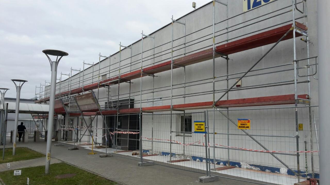 Budynek w trakcie remontu z rozstawionym rusztowaniem na całej długości elewacji, zabezpieczony siatką i tablicami ostrzegawczymi.