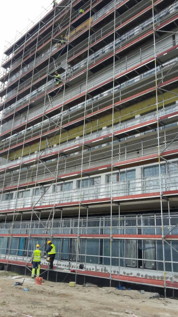 Wielopiętrowy budynek w trakcie remontu, otoczony rusztowaniem. Dwóch pracowników w żółtych kamizelkach na rusztowaniu przy oknach, inni na wyższych poziomach. Widoczne materiały budowlane.