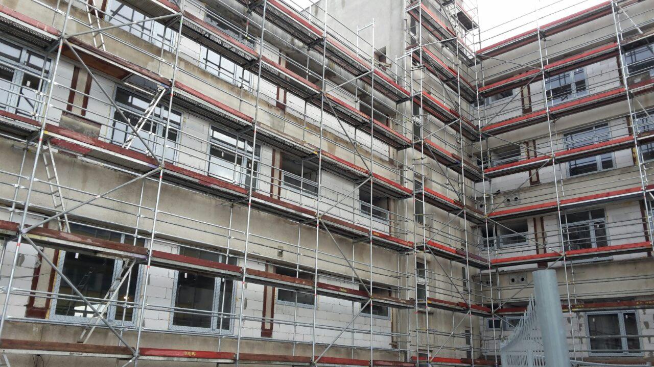 Budynek wielopiętrowy w trakcie remontu, otoczony rusztowaniami z czerwonymi elementami zabezpieczającymi, widoczne okna i ściany z betonu komórkowego.