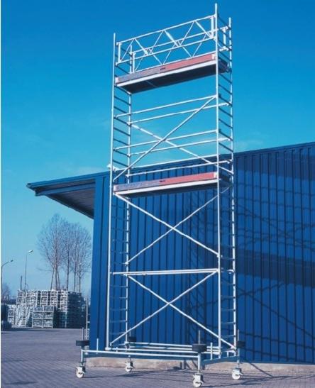 Wysokie, metalowe rusztowanie na kółkach, ustawione na betonowej nawierzchni, z niebieskim budynkiem w tle i stosem palet w oddali.