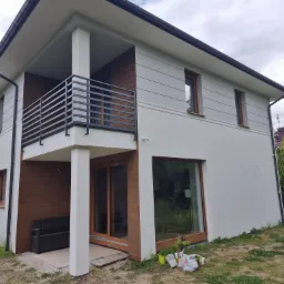 Dwukondygnacyjny dom z wykończoną elewacją w odcieniach bieli i szarości, z elementami drewnianymi, balkonem z czarną balustradą i dużymi oknami, otoczony zielenią.