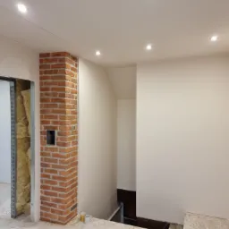 Wnętrze w trakcie prac wykończeniowych: fragment ściany z cegły klinkierowej, ocieplenie wełną mineralną widoczne w otworze drzwiowym, sufit z oświetleniem punktowym, świeżo pomalowana ściana.