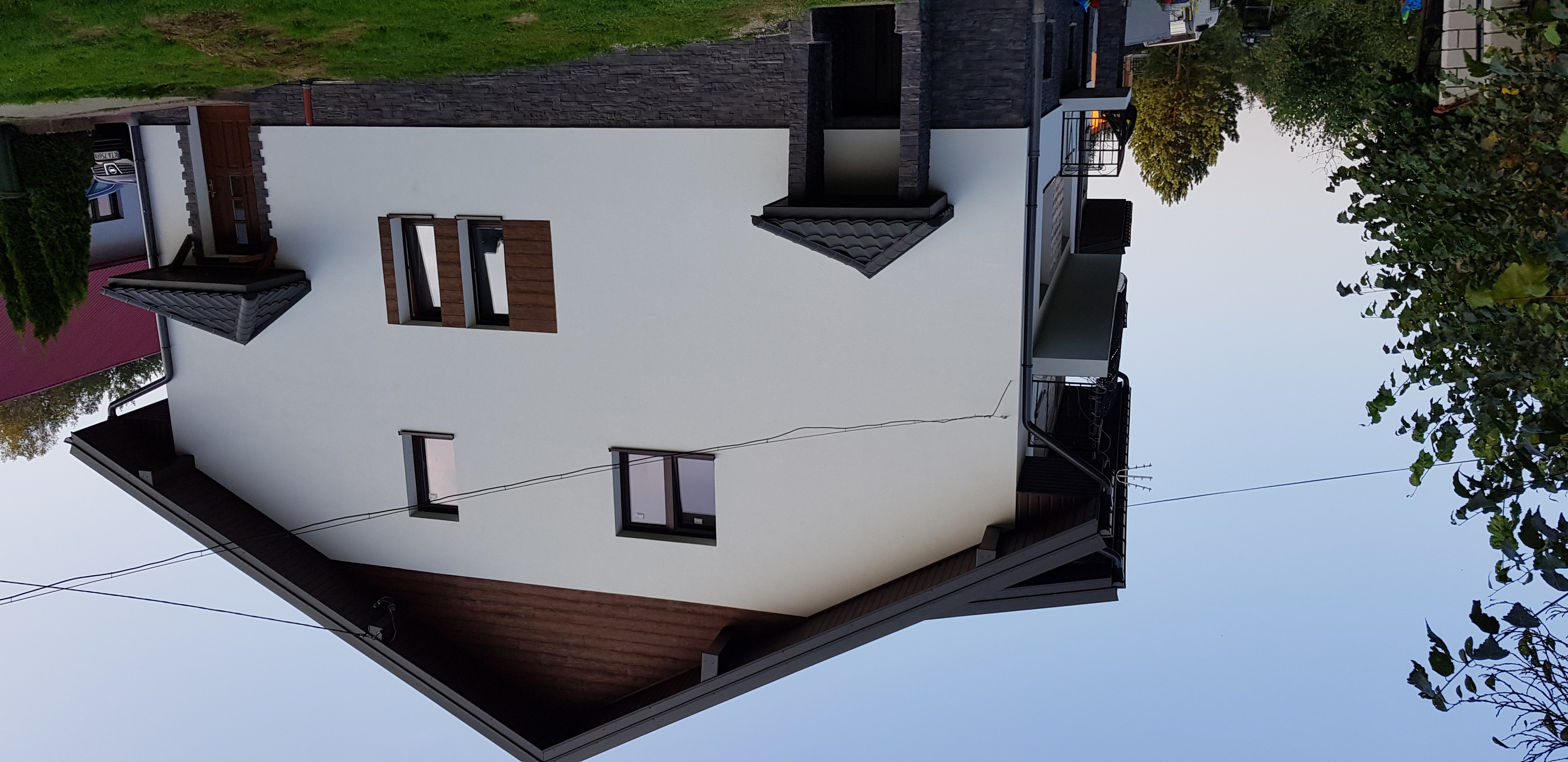 Biała elewacja domu jednorodzinnego z brązowymi oknami i grafitowym dachem, widok z dołu na tle błękitnego nieba.