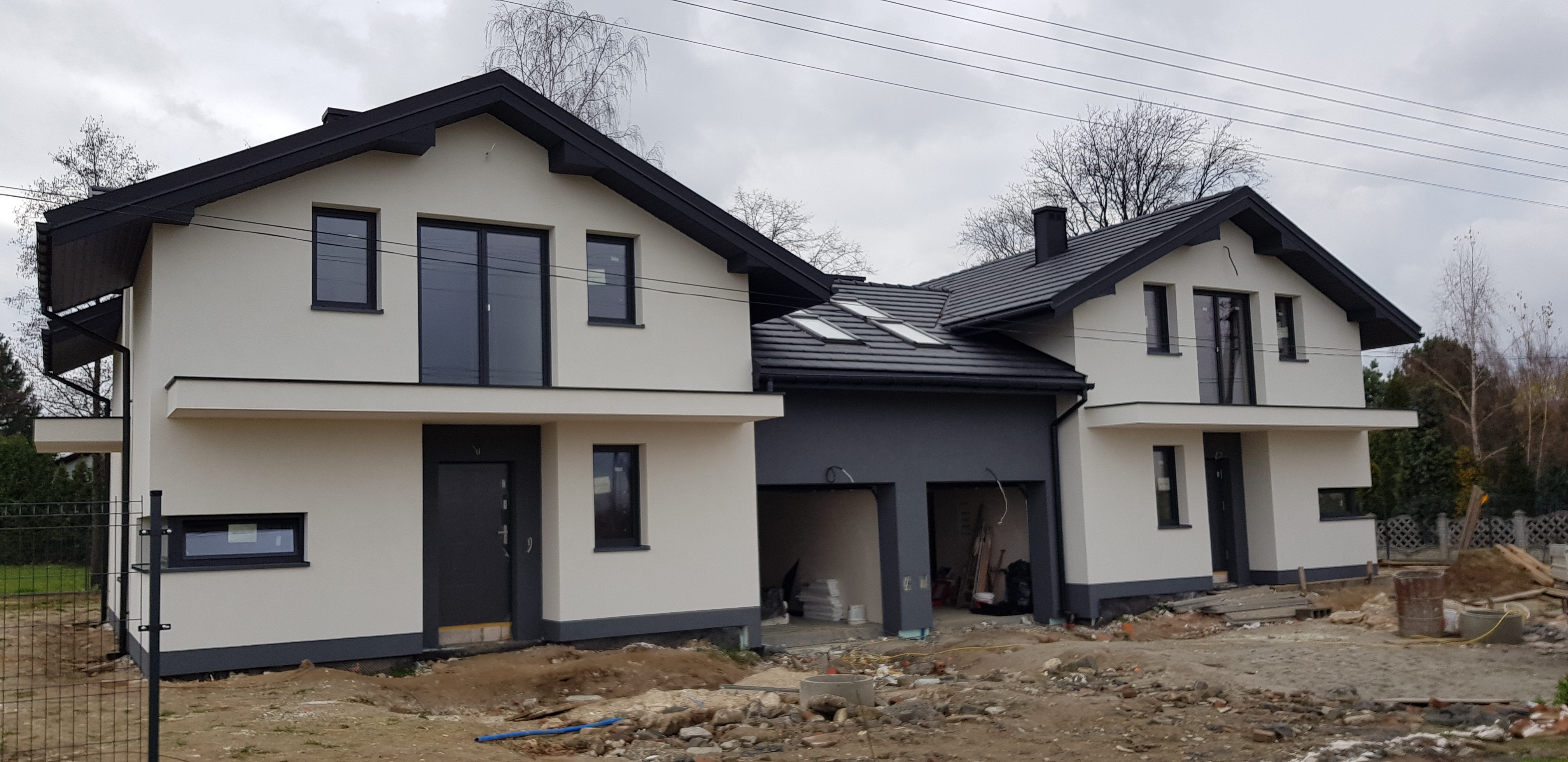 Dwa nowoczesne, dwukondygnacyjne domy jednorodzinne z płaskimi dachami i czarnymi ramami okiennymi, połączone garażem z dwoma bramami, w trakcie prac wykończeniowych elewacji zewnętrznej, widoczne...