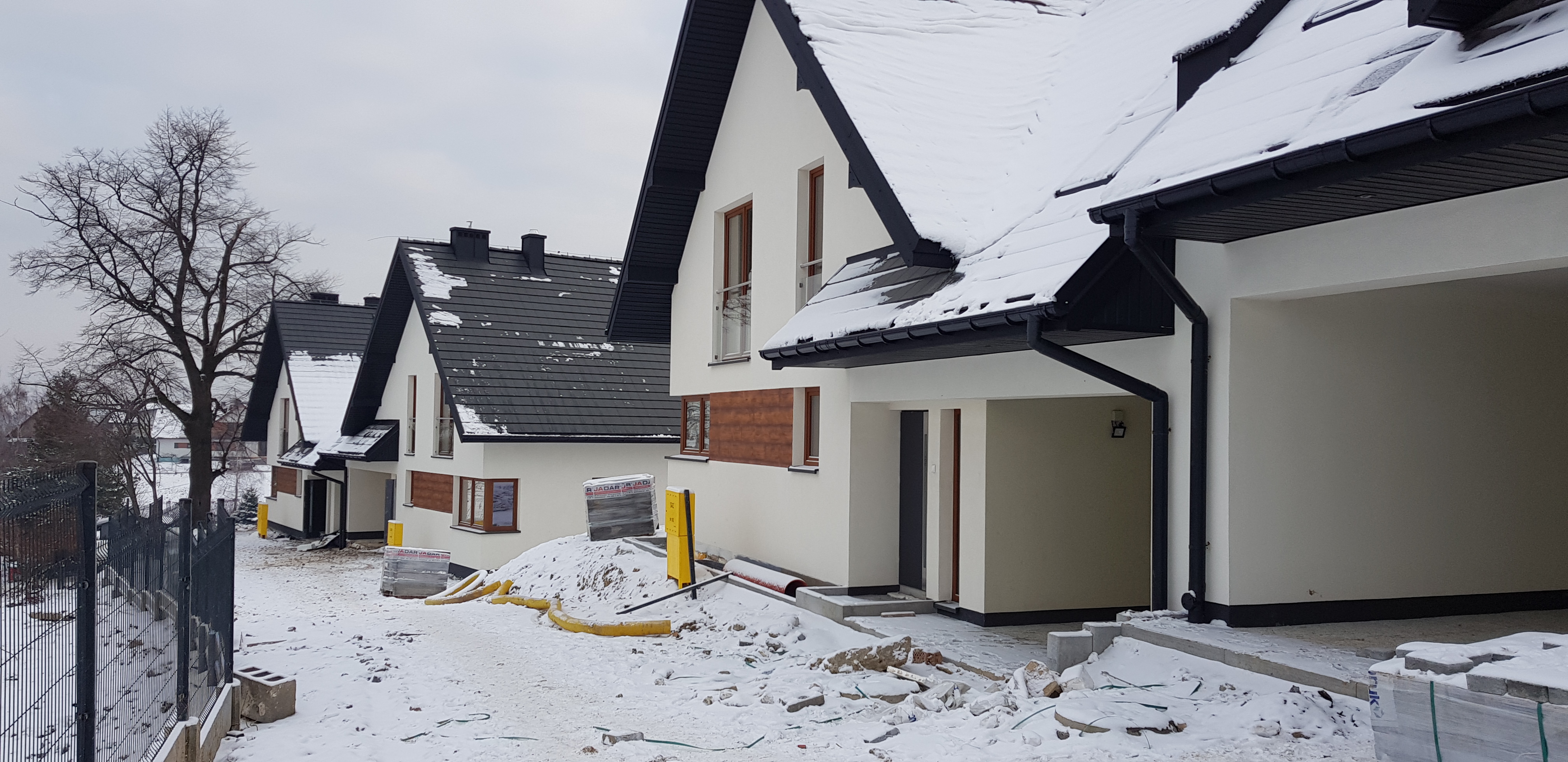 Rząd nowych, ośnieżonych domów jednorodzinnych z białymi elewacjami i czarnymi dachami, widok z zewnątrz, w tle gołe drzewo i ogrodzenie.