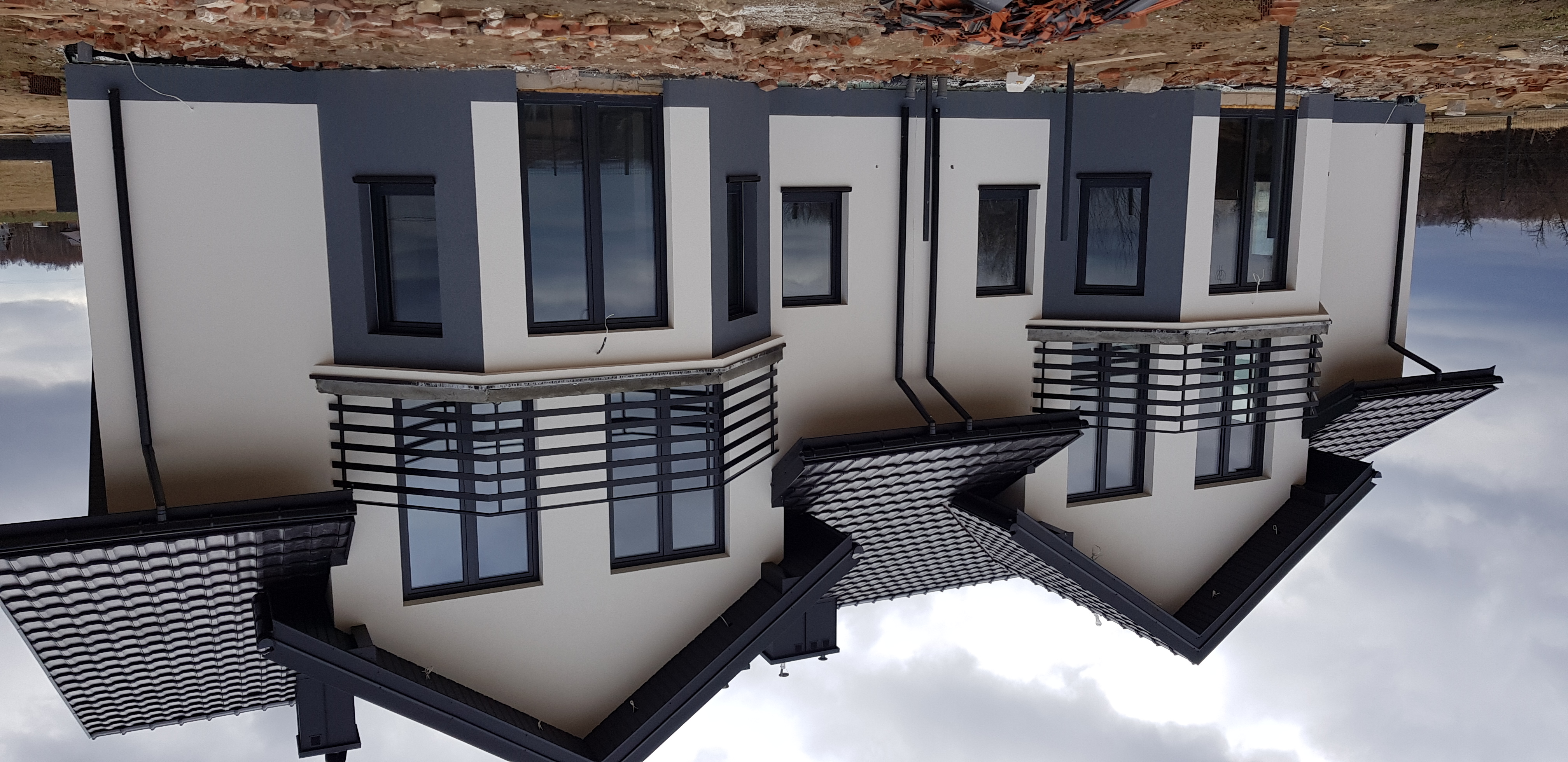 Dwukondygnacyjny dom z elewacją w odcieniach szarości i bieli, z ciemną dachówką i czarnymi ramami okiennymi, widziany z zewnątrz w pochmurny dzień.