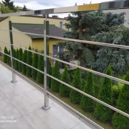 Błyszcząca, metalowa balustrada z poziomymi prętami, zamontowana na balkonie z jasnymi płytkami. W tle rząd małych drzewek i budynek.