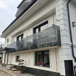 Nowoczesna balustrada szklana z metalowymi mocowaniami na balkonie domu z białą elewacją. Widok z dołu, odbicie drzew w szkle.