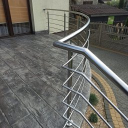 JURA-TECH - Błyszcząca, nierdzewna balustrada balkonowa z zaokrąglonym profilem, zamontowana na balkonie z betonową podłogą imitującą drewno. Widok na posesję.