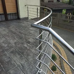 Błyszcząca, nierdzewna balustrada balkonowa z zaokrąglonym profilem, zamontowana na balkonie z betonową podłogą imitującą drewno. Widok na posesję.
