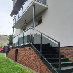 JURA-TECH - Szklane balustrady na balkonie i schodach zewnętrznych domu z ceglaną podmurówką, widok z dołu. Minimalistyczny design, szare ramy i przezroczyste szkło.