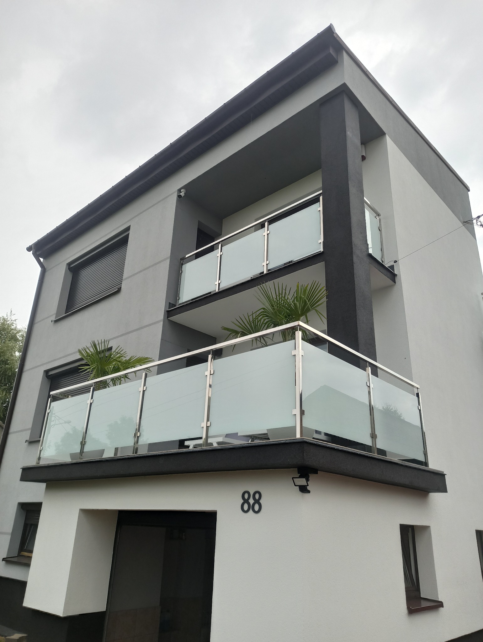 Dwupiętrowy dom z szarymi ścianami i balkonami z balustradami szklanymi i stalowymi. Na balkonach widoczne rośliny. Numer domu 88.