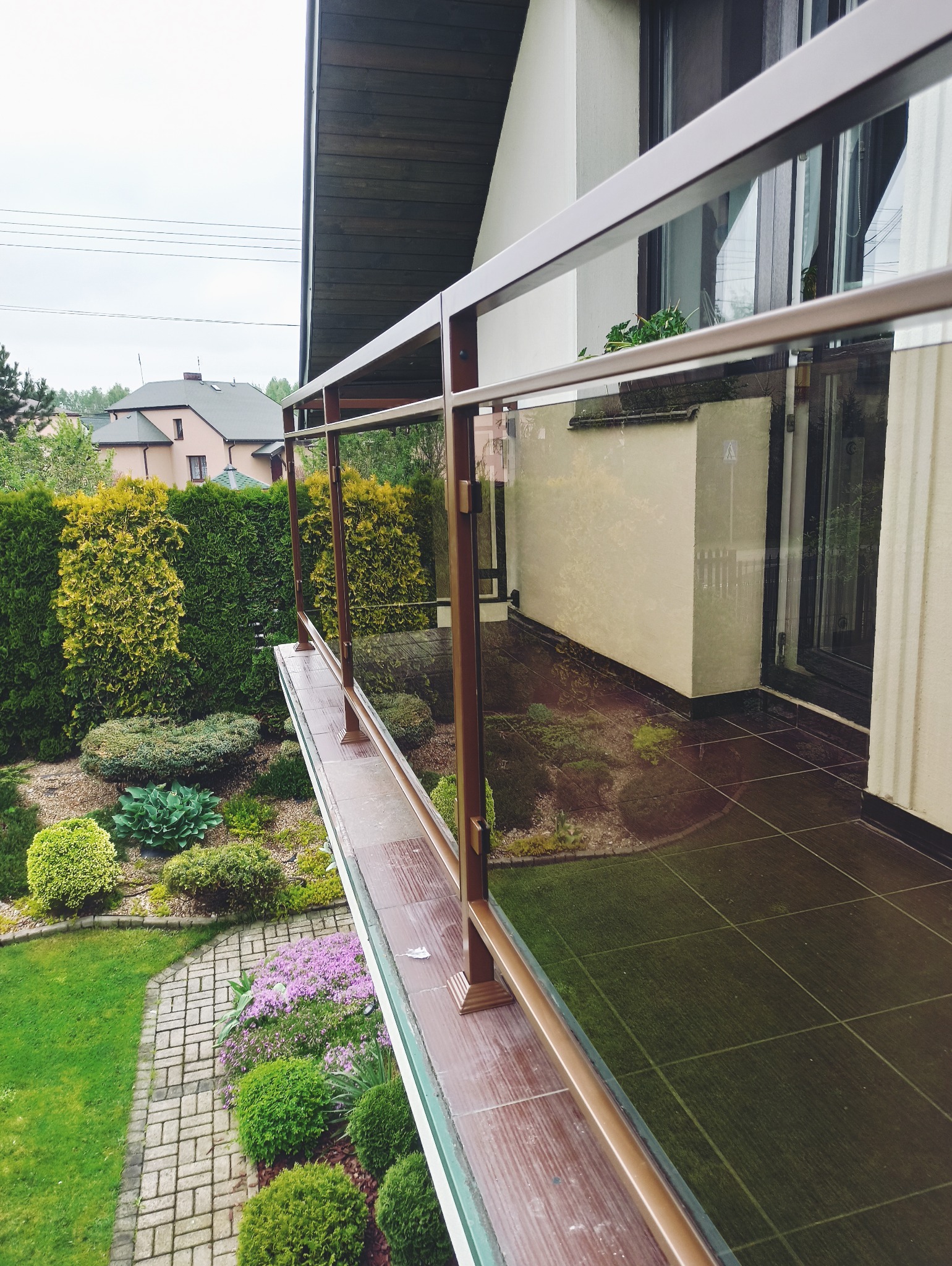 Balkon z brązową, szklaną balustradą, z widokiem na zadbany ogród i fragment domu. Ujęcie z góry, podkreślające estetykę wykonania.