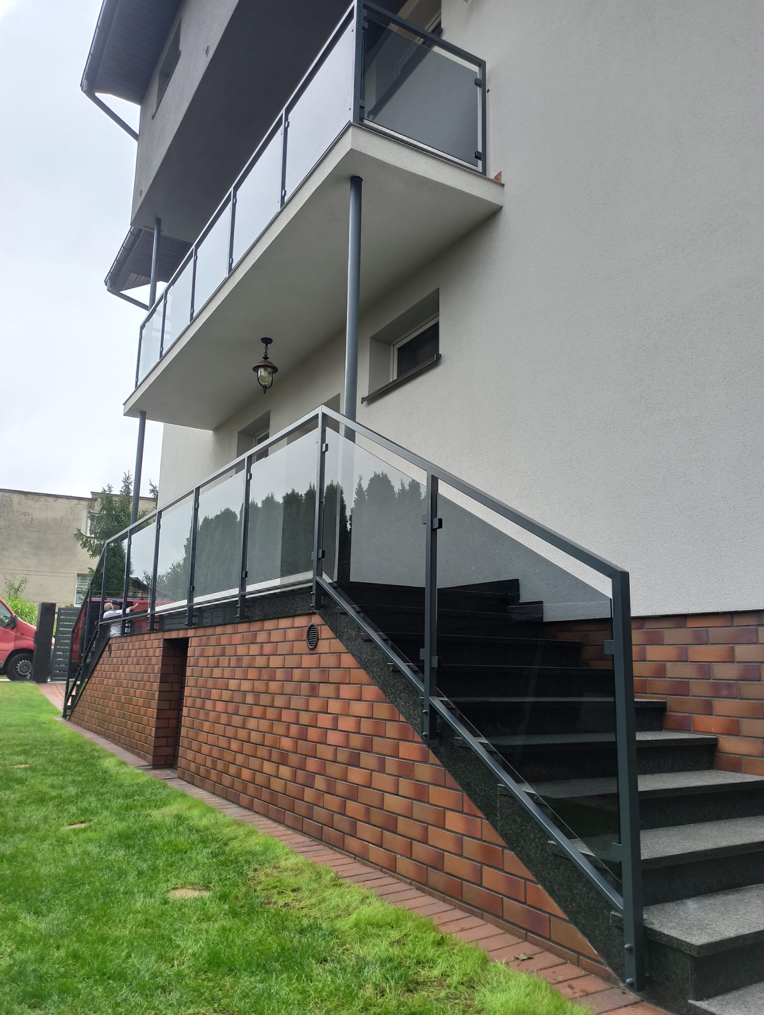 Szklane balustrady na balkonie i schodach zewnętrznych domu z ceglaną podmurówką, widok z dołu. Minimalistyczny design, szare ramy i przezroczyste szkło.