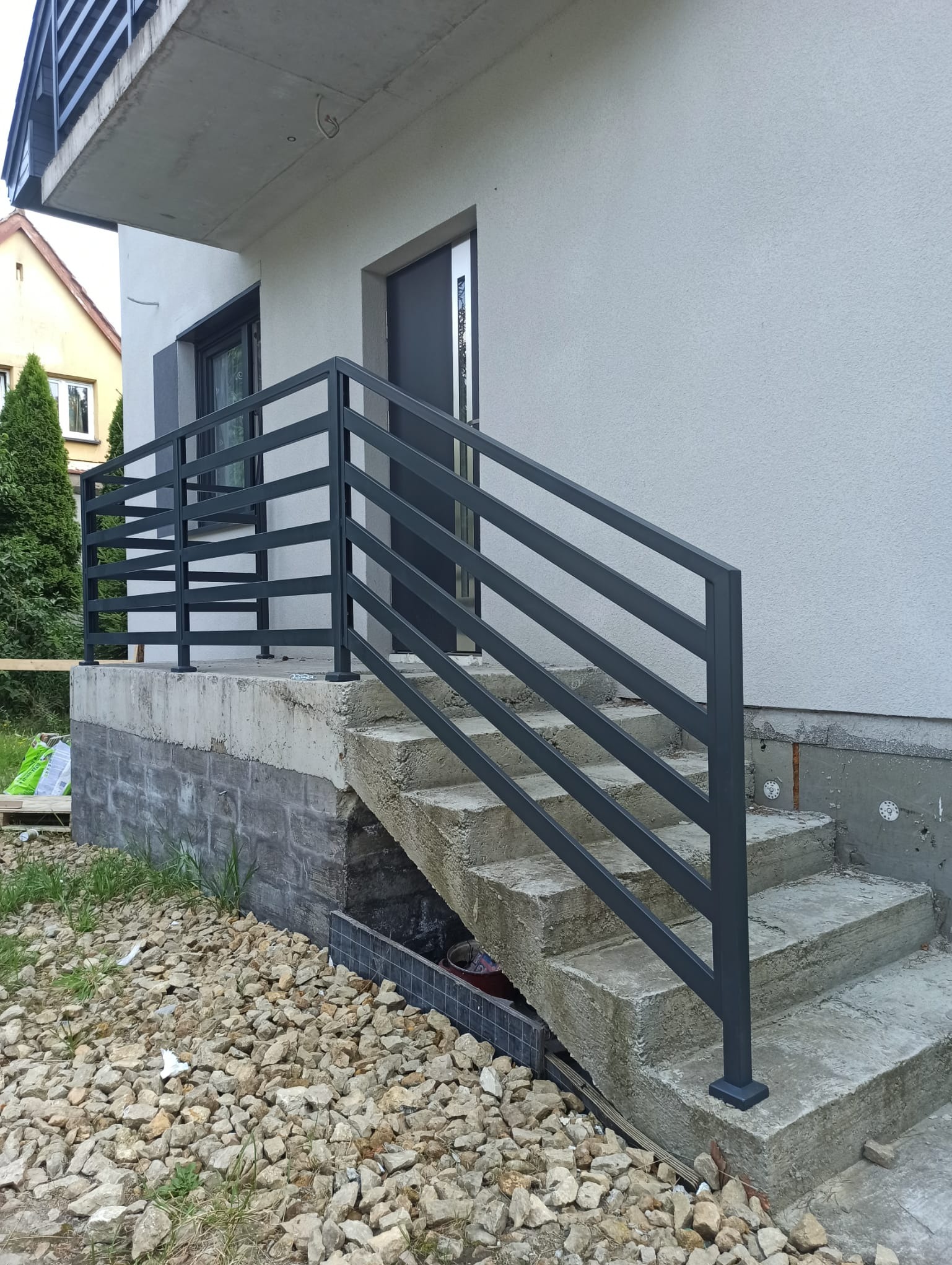 Zewnętrzne schody z szarą, metalową balustradą o minimalistycznym designie, prowadzące do drzwi wejściowych nowoczesnego domu o jasnej elewacji.