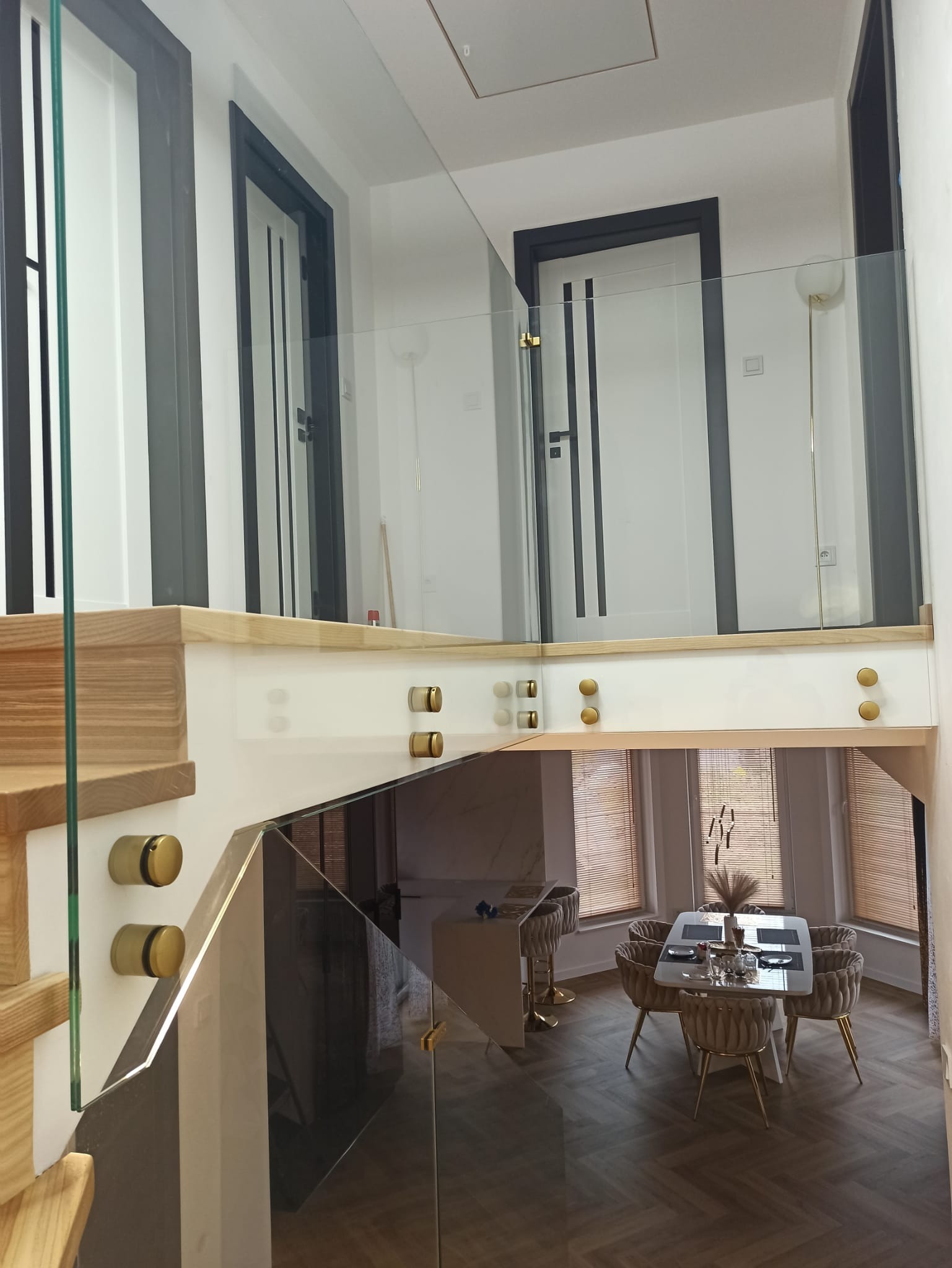 Szklana balustrada schodowa z drewnianymi stopniami i złotymi mocowaniami, widok z góry na jadalnię w jasnym wnętrzu.