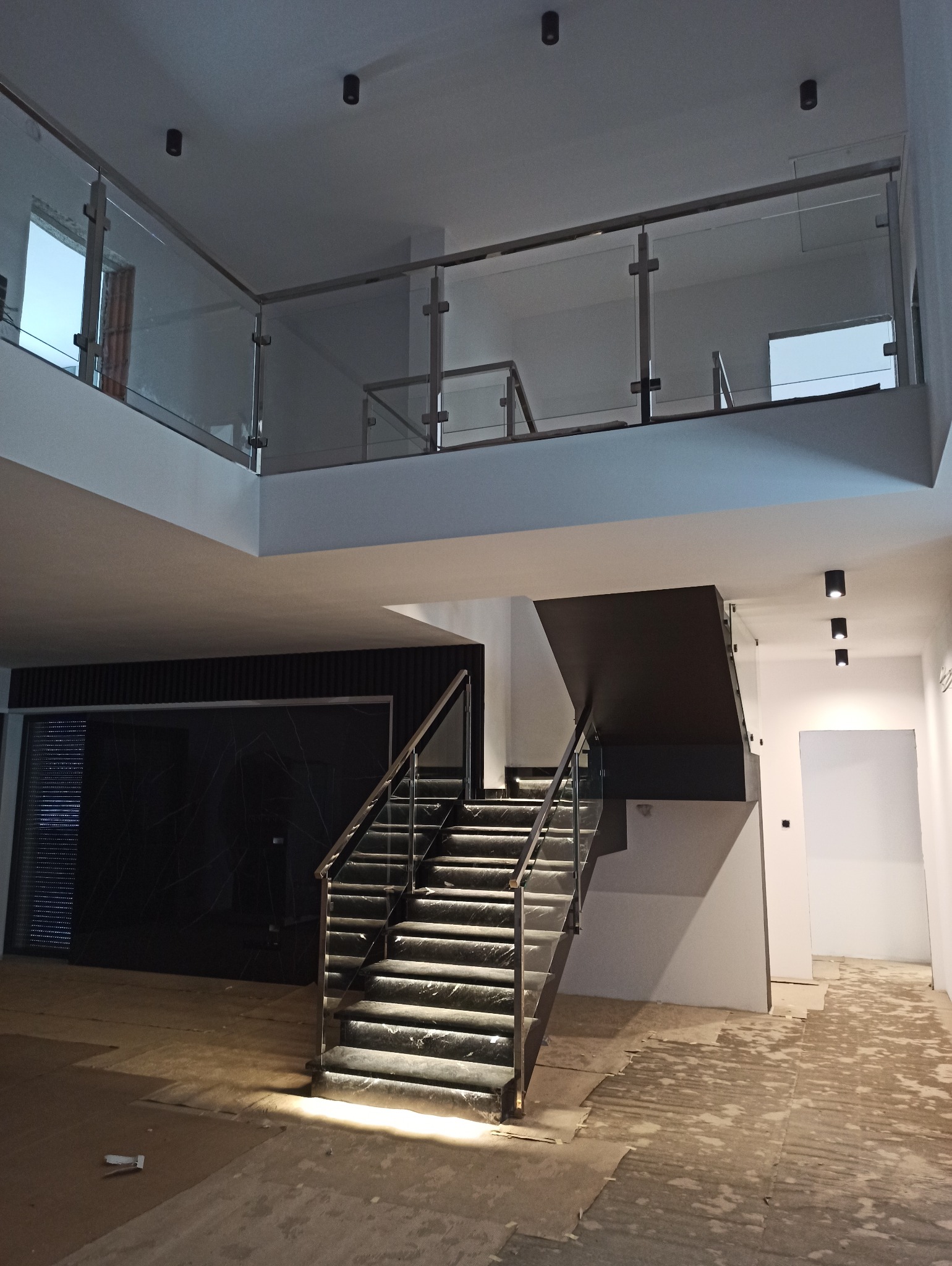 Dwupoziomowe wnętrze z nowoczesnymi schodami i szklanymi balustradami. Widoczne stalowe elementy konstrukcyjne i marmurowe stopnie. Oświetlenie punktowe.