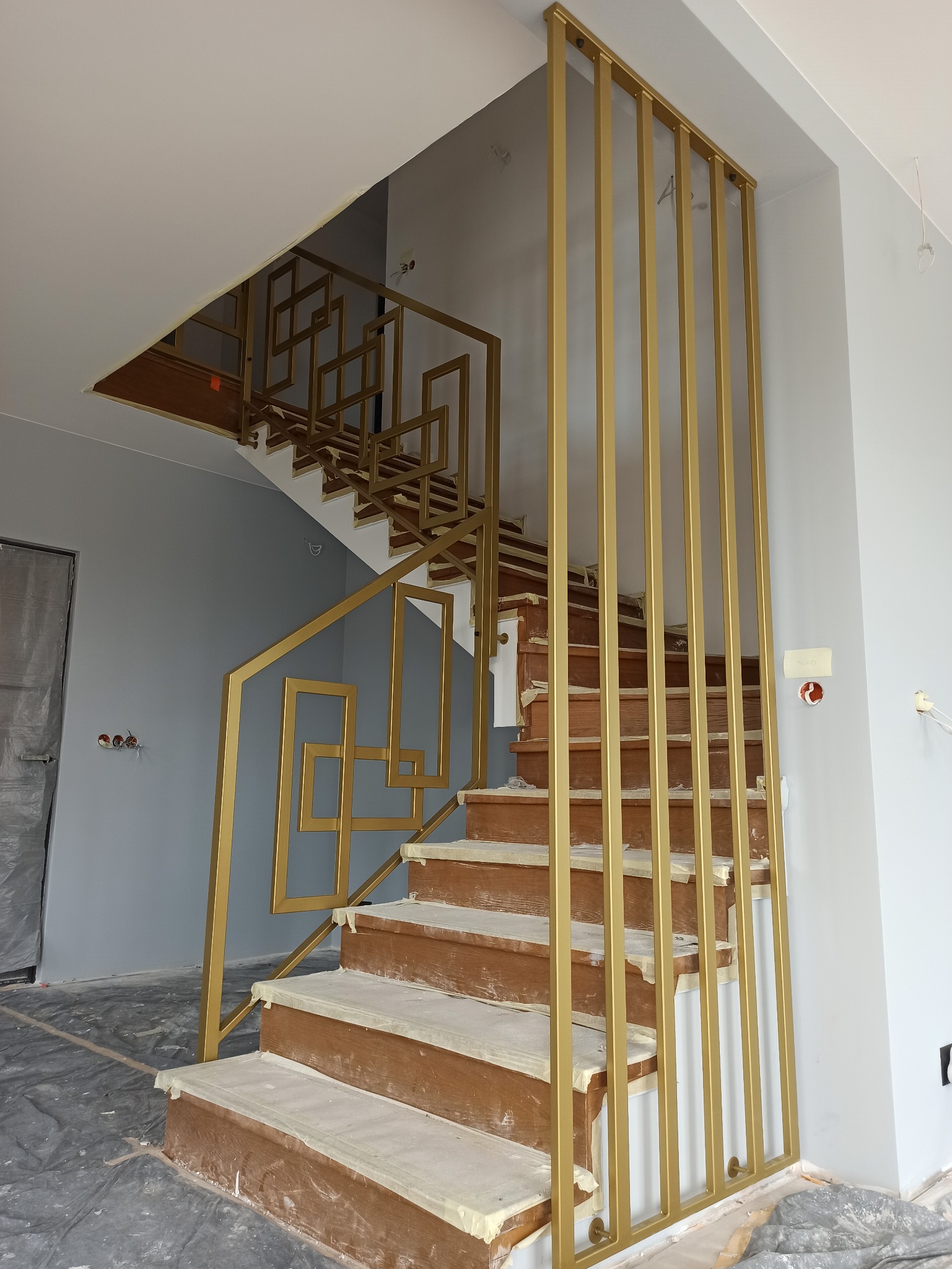 Złote, geometryczne balustrady schodowe w trakcie montażu. Widoczne zabezpieczenia na stopniach i podłodze. Nowoczesny design, pionowe elementy z metalu.