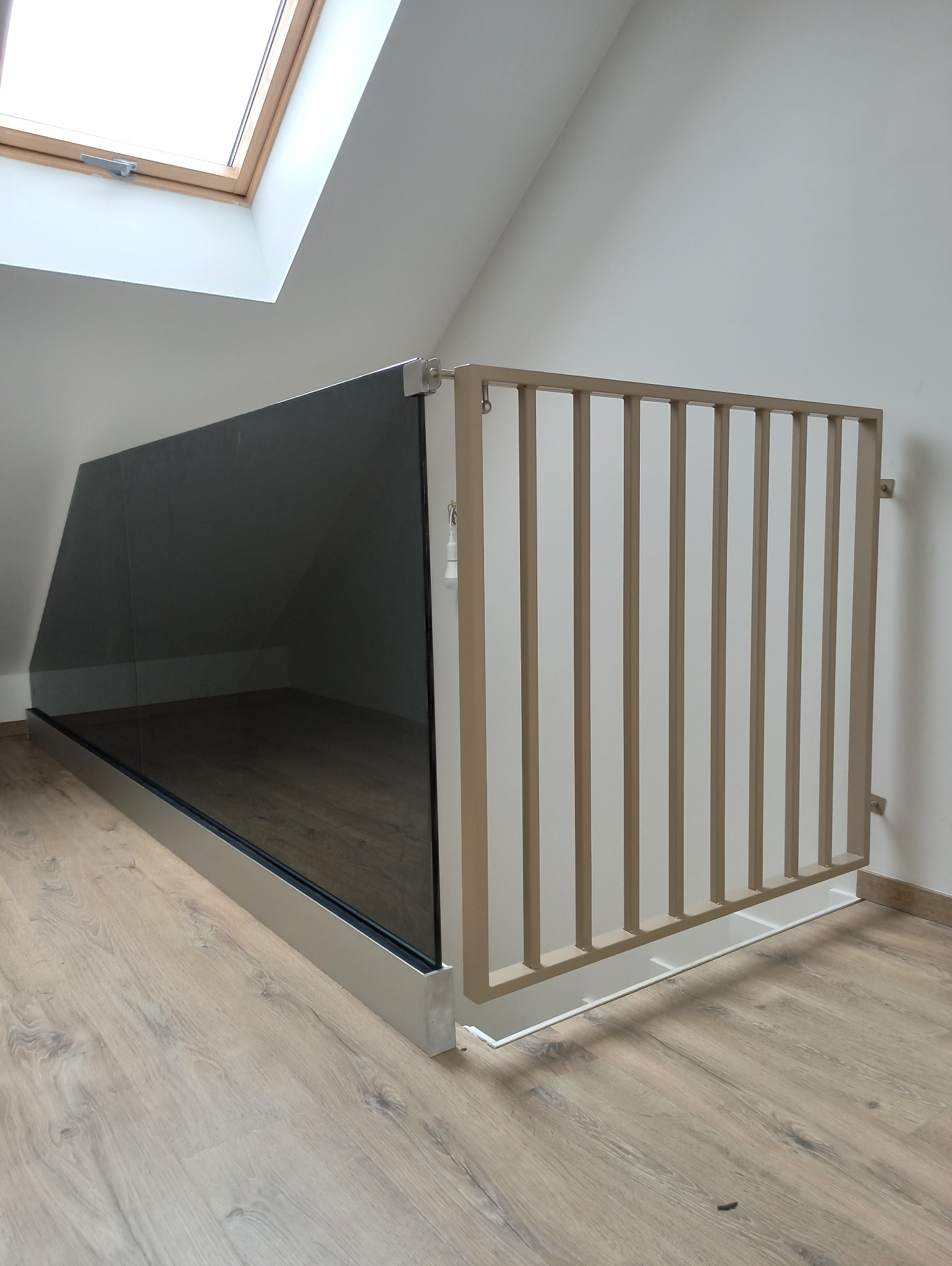Szklana balustrada z drewnianymi szczeblami zabezpieczająca antresolę na poddaszu, widok od strony podłogi, oświetlenie naturalne przez okno dachowe.