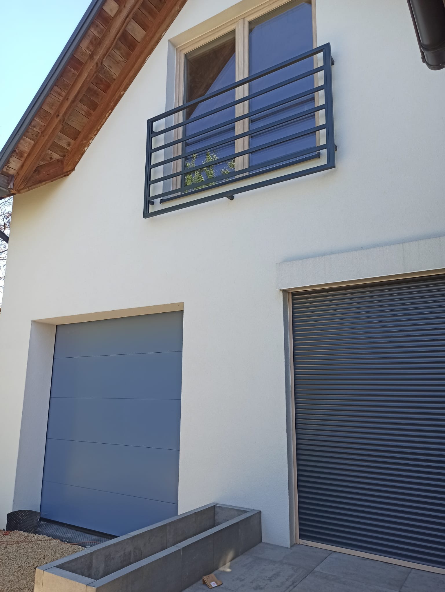 Minimalistyczna, czarna balustrada okienna z poziomych prętów na tle białej fasady domu. Widoczne dwie bramy garażowe.