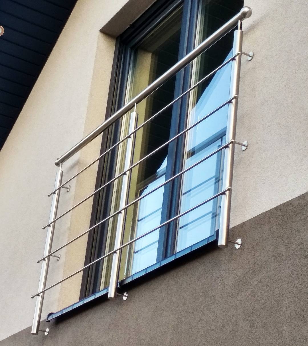 Zewnętrzna balustrada szklana z elementami ze stali nierdzewnej, zamocowana do elewacji budynku. Widok z dołu, na tle okna.