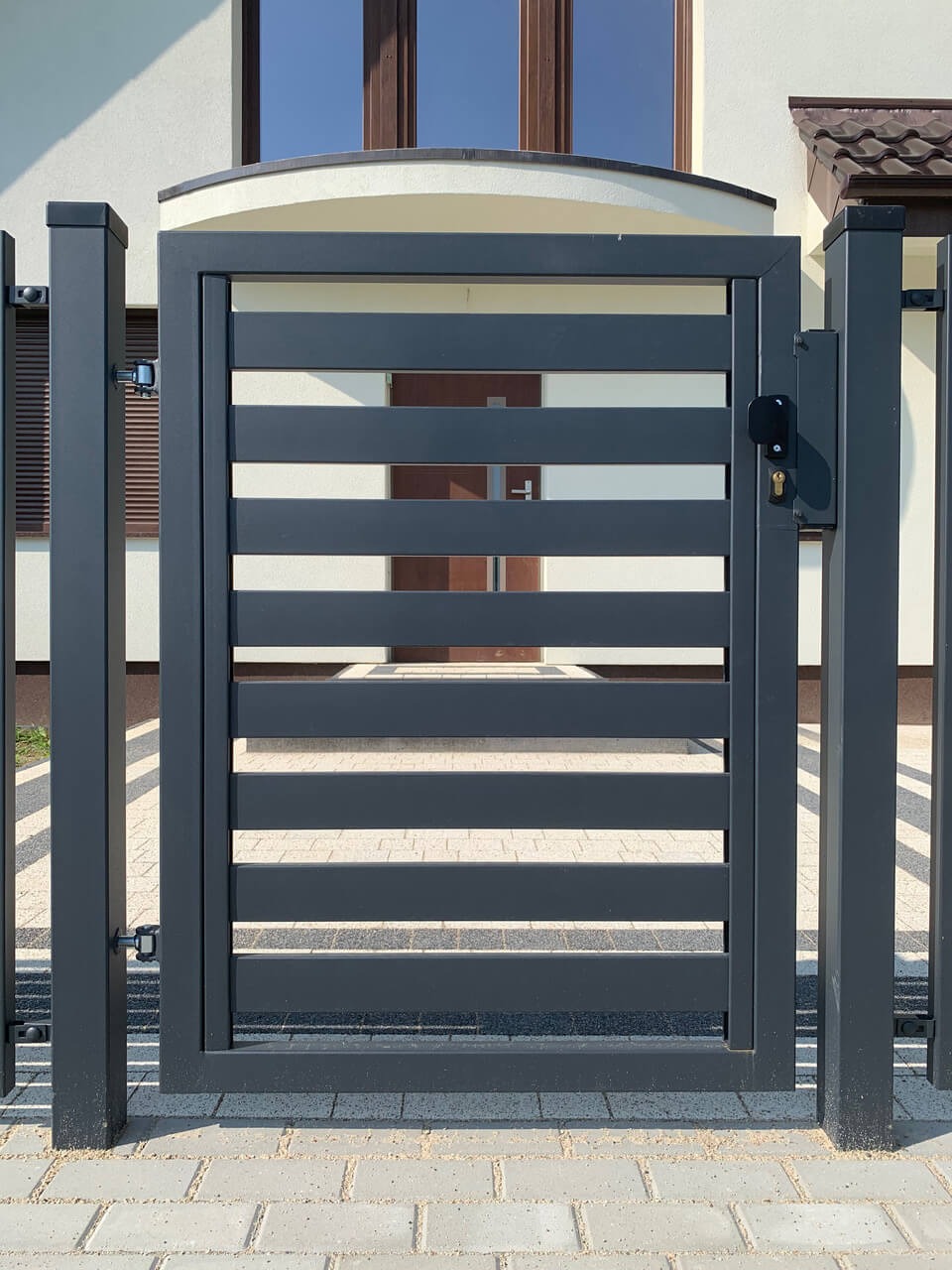 Nowoczesna, metalowa furtka w kolorze grafitowym z poziomymi szczeblami, zamontowana przy posesji z jasną elewacją i brukowanym podjazdem. Minimalistyczny design.