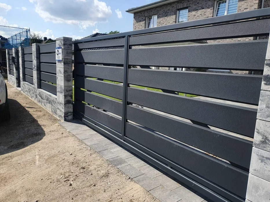 Nowoczesna, przesuwna brama w kolorze grafitowym z poziomymi panelami, osadzona na słupkach z szarej kostki. Widok na posesję z zewnątrz.