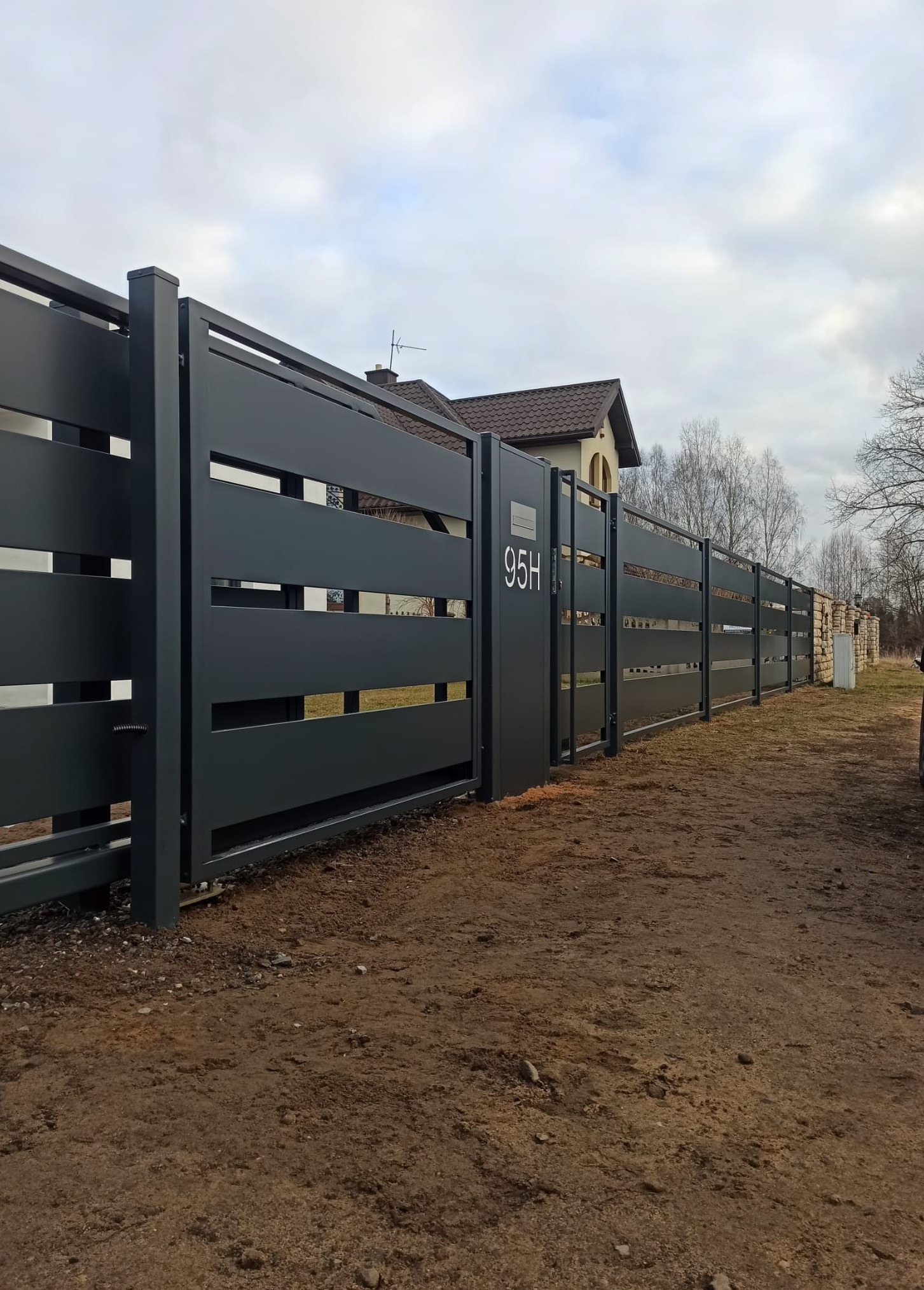 Nowoczesna, pozioma, metalowa brama przesuwna w kolorze antracytu z numerem 95H. W tle widoczny dom i fragment ogrodzenia z kamienia.