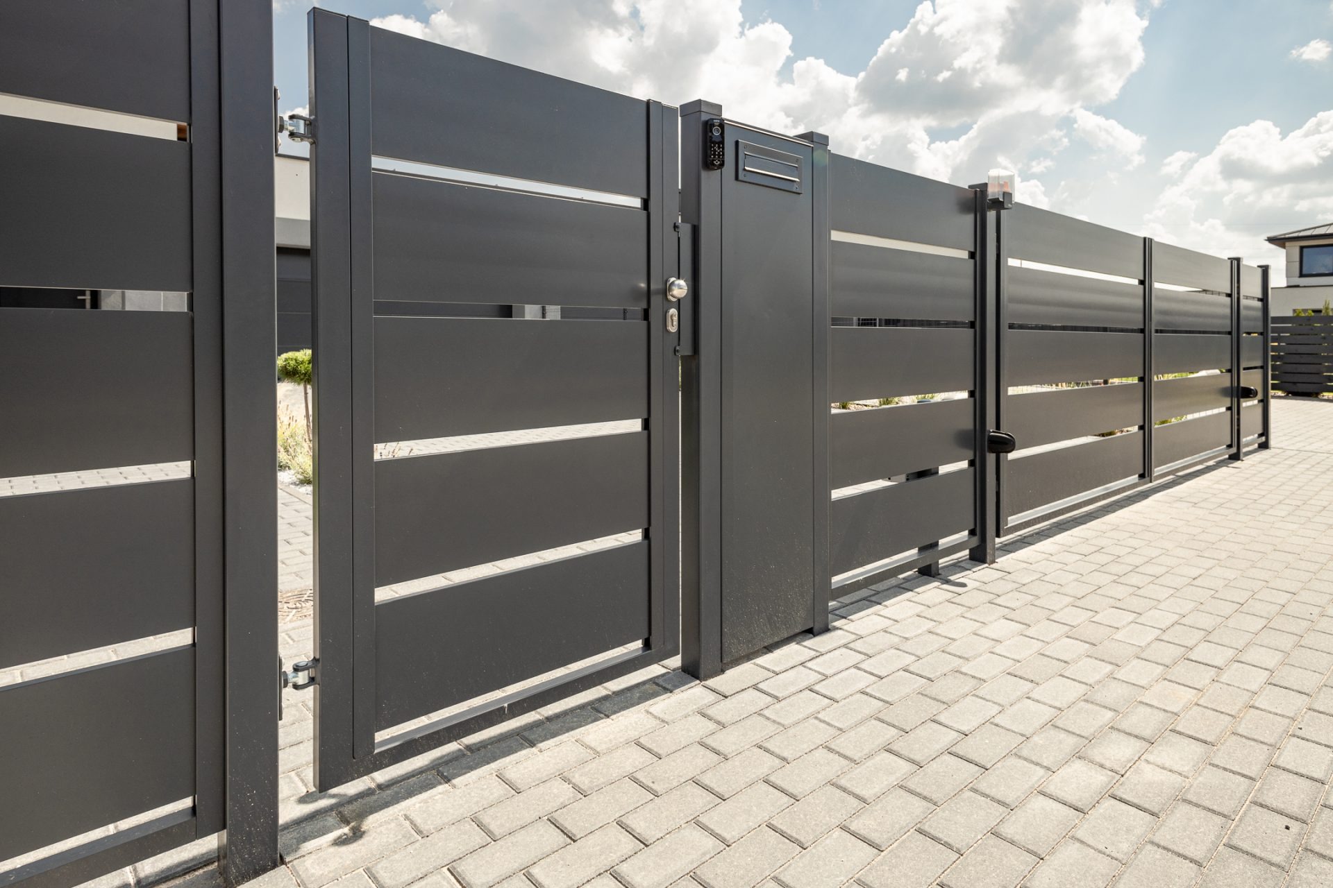 Nowoczesna, grafitowa brama wjazdowa z poziomymi szczeblami i domofonem, zamontowana na posesji z brukowanym podjazdem w słoneczny dzień.