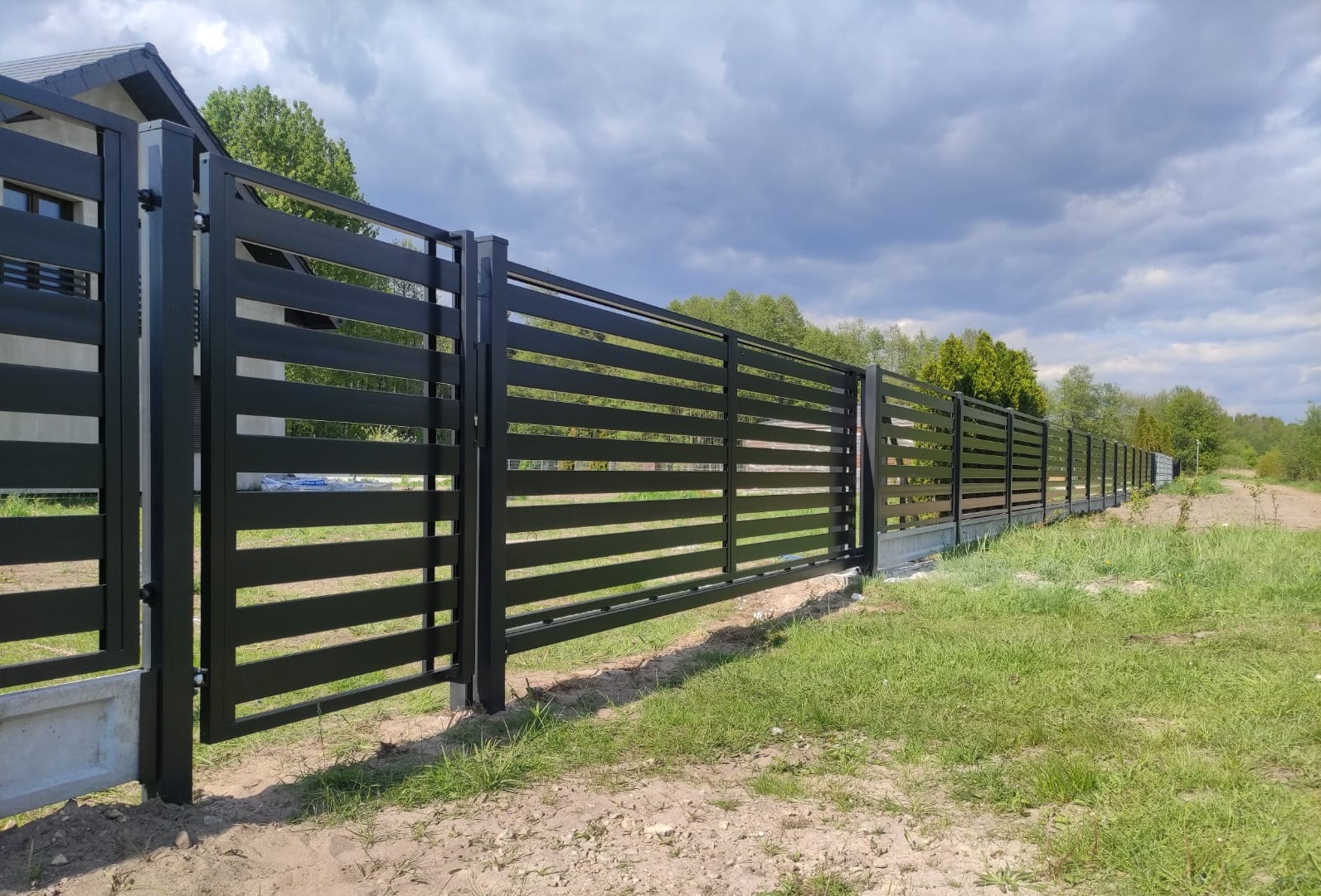 Nowoczesne, poziome przęsła czarnego ogrodzenia panelowego na betonowej podmurówce, z widoczną furtką i fragmentem domu w tle.