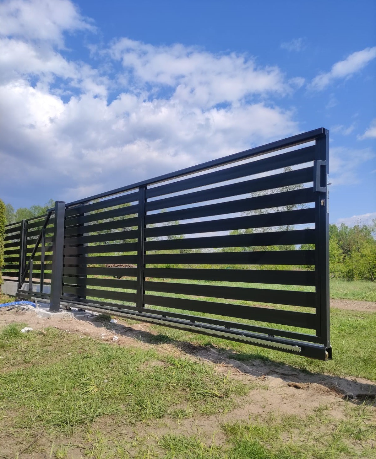 Nowoczesna, przesuwna brama posesyjna z poziomych, czarnych profili, na tle błękitnego nieba z chmurami. Widoczny mechanizm jezdny bramy.
