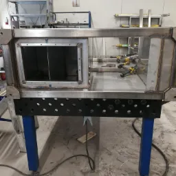 Konstrukcja ze stali spawana z profili zamkniętych, na niebieskich nogach, z widocznymi śladami spawania i mocowania elementów, w tle regały warsztatowe.