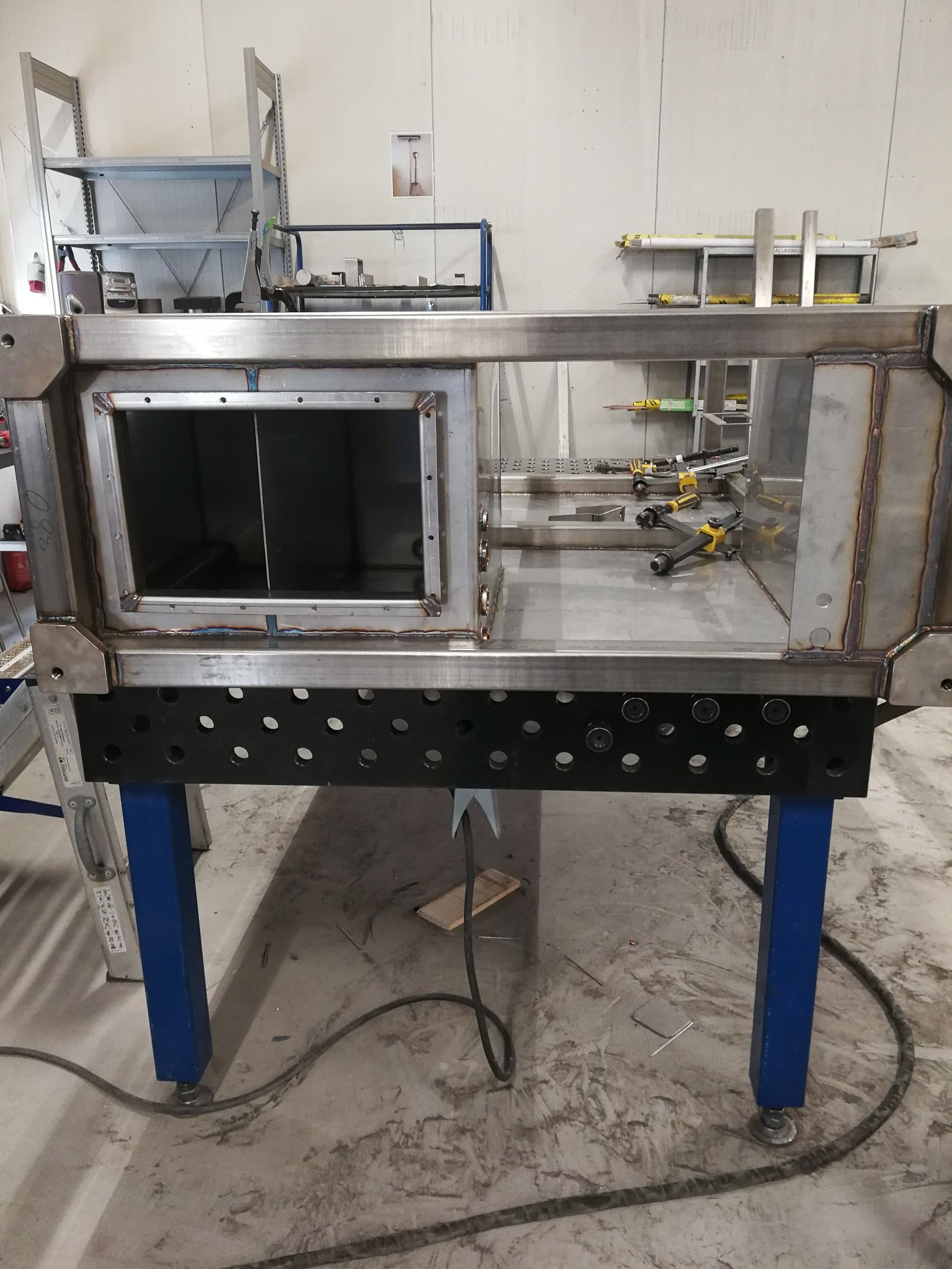 Konstrukcja ze stali spawana z profili zamkniętych, na niebieskich nogach, z widocznymi śladami spawania i mocowania elementów, w tle regały warsztatowe.