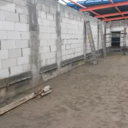 Wznoszenie ścian z betonu komórkowego na fundamencie z widocznymi poziomami zbrojenia, w tle konstrukcja stalowa dachu i drabina oparta o ścianę.