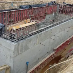 Wykop pod fundamenty z szalunkiem i zbrojeniem, widoczne elementy konstrukcyjne oraz narzędzia na placu budowy.