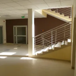 Wnętrze budynku w stanie deweloperskim: widok na klatkę schodową z metalową balustradą, drzwi wejściowe i sufit podwieszany z oświetleniem.