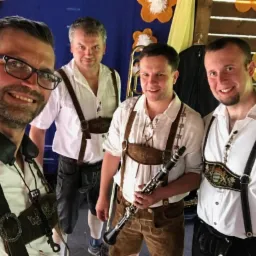 Czterech mężczyzn w bawarskich strojach ludowych pozuje do selfie, jeden trzyma klarnet, w tle dekoracje z kwiatów.
