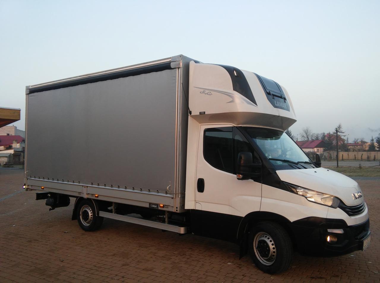 Biało-czarny bus dostawczy z szarą plandeką na tle brukowanej powierzchni i budynków w oddali.