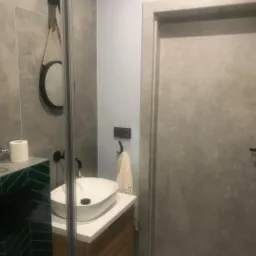 Wykończona łazienka z umywalką nablatową, szafką z drewnianym frontem, lustrem na sznurze i szarymi płytkami imitującymi beton.