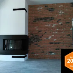 Nowoczesny kominek narożny z czarną obudową, ceglana ściana w tle z widocznymi fragmentami białej farby, na podłodze szare płytki, w prawym dolnym rogu logo Oferteo Najlepsi 2017.