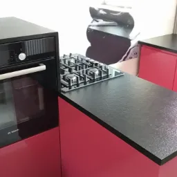 Nowoczesna kuchnia z czarnym blatem z kamienia, zabudowanym piekarnikiem Bosch i płytą gazową, r&oacute;żowe szafki kuchenne, minimalistyczny design.