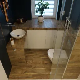 Nowoczesna łazienka z drewnopodobnymi płytkami, szklaną kabiną prysznicową, umywalką nablatową i rośliną w doniczce na blacie przy oknie.