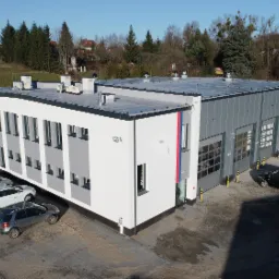 Nowoczesna hala z szarymi segmentowymi bramami garażowymi, białymi ścianami z szarymi akcentami i pasem w kolorach czerwonym i niebieskim, na parkingu stoi kilka samochodów osobowych i dostawczy...