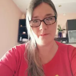 Portret kobiety w okularach, w czerwonej koszulce z cekinami, na tle jasnej ściany i ciemnych półek z dekoracjami.