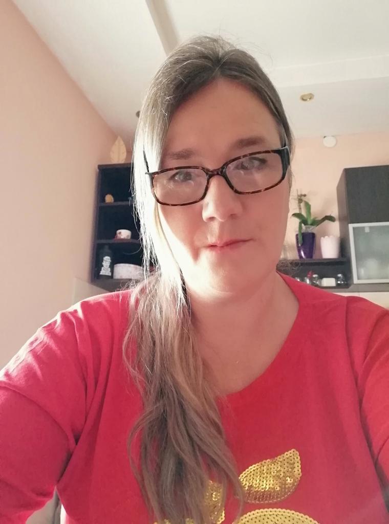 Portret kobiety w okularach, w czerwonej koszulce z cekinami, na tle jasnej ściany i ciemnych półek z dekoracjami.