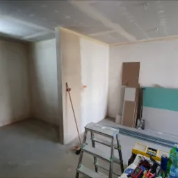 Wnętrze w trakcie remontu: ściany częściowo pokryte płytami gipsowo-kartonowymi, widoczne narzędzia, drabina aluminiowa, profile metalowe i materiały budowlane, surowy beton na podłodze...