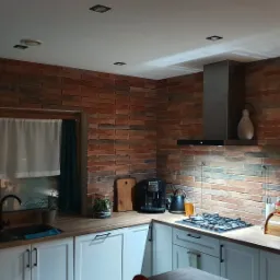 Kuchnia z ceglaną ścianą, białymi szafkami i drewnianym blatem, oświetlona punktowymi lampami sufitowymi, z widocznym ekspresem do kawy, płytą gazową i okapem.