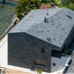 Nowoczesny dom jednorodzinny z dachem pokrytym ciemnoszarą dachówką łupkową, widok z góry na tle jeziora.